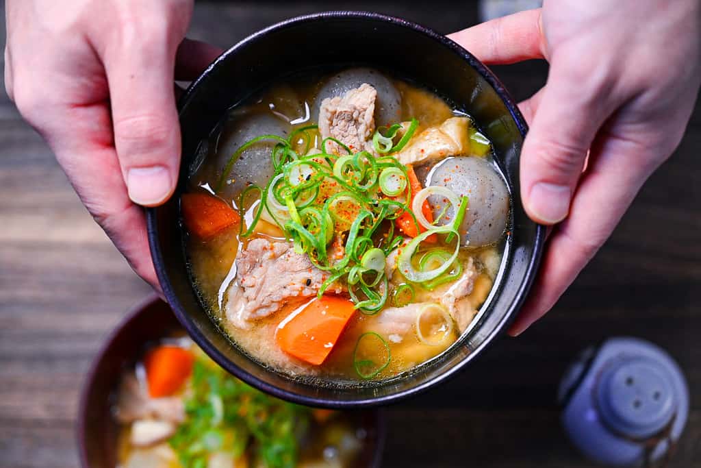 Two hands holding a black bowl of tonjiru (pork and vegetable miso soup)