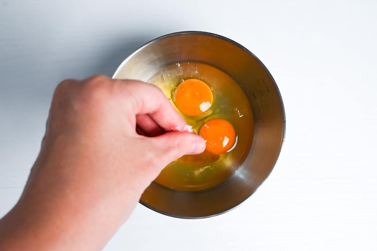 adding a pinch of salt to eggs in a metal mixing bowl