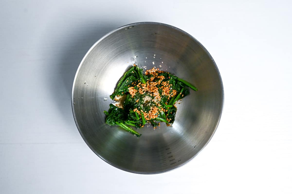 mixing blanched spinach with sesame oil and ground sesame in a metal mixing bowl