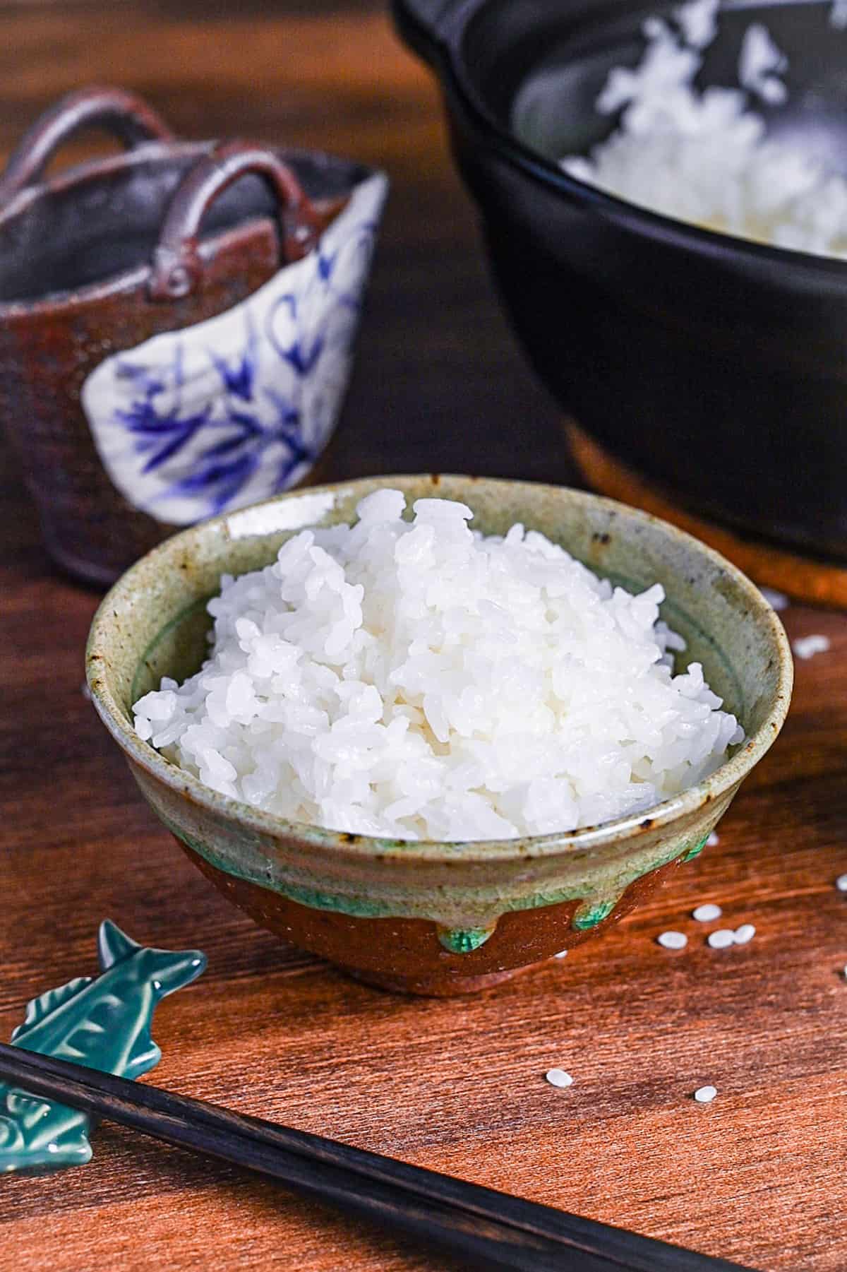 Stove-cooked Japanese white rice in a brown and green ceramic bowl next to a cooking pot, rice paddle cup and black chopsticks