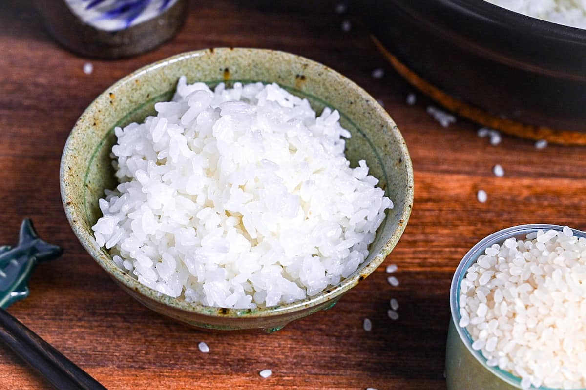 Stove-cooked Japanese white rice in a brown and green ceramic bowl next to a cooking pot, rice paddle cup and black chopsticks