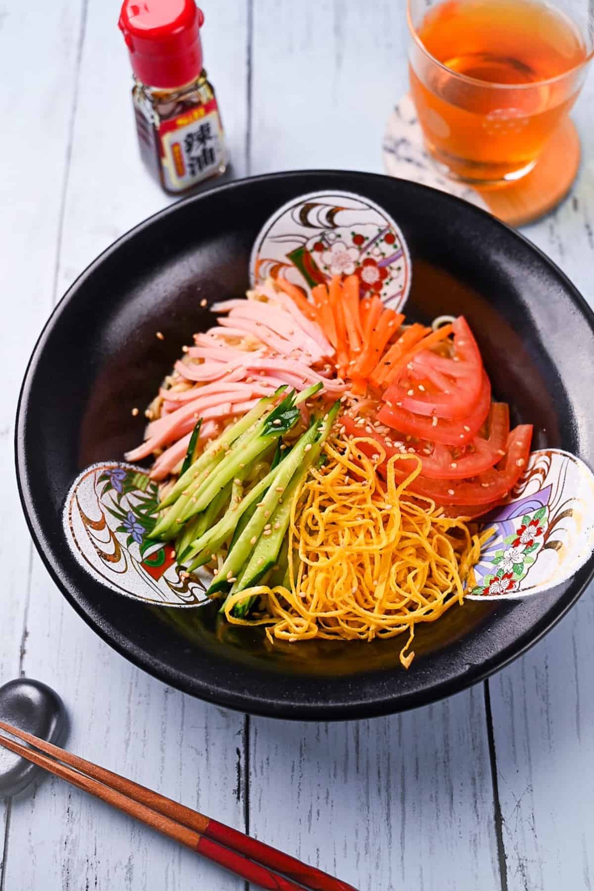 Hiyashi chuka made with ramen noodles topped with ham, carrots, tomatoes, egg crepe and cucumber with a tangy soy-based sauce in a black Chinese style bowl
