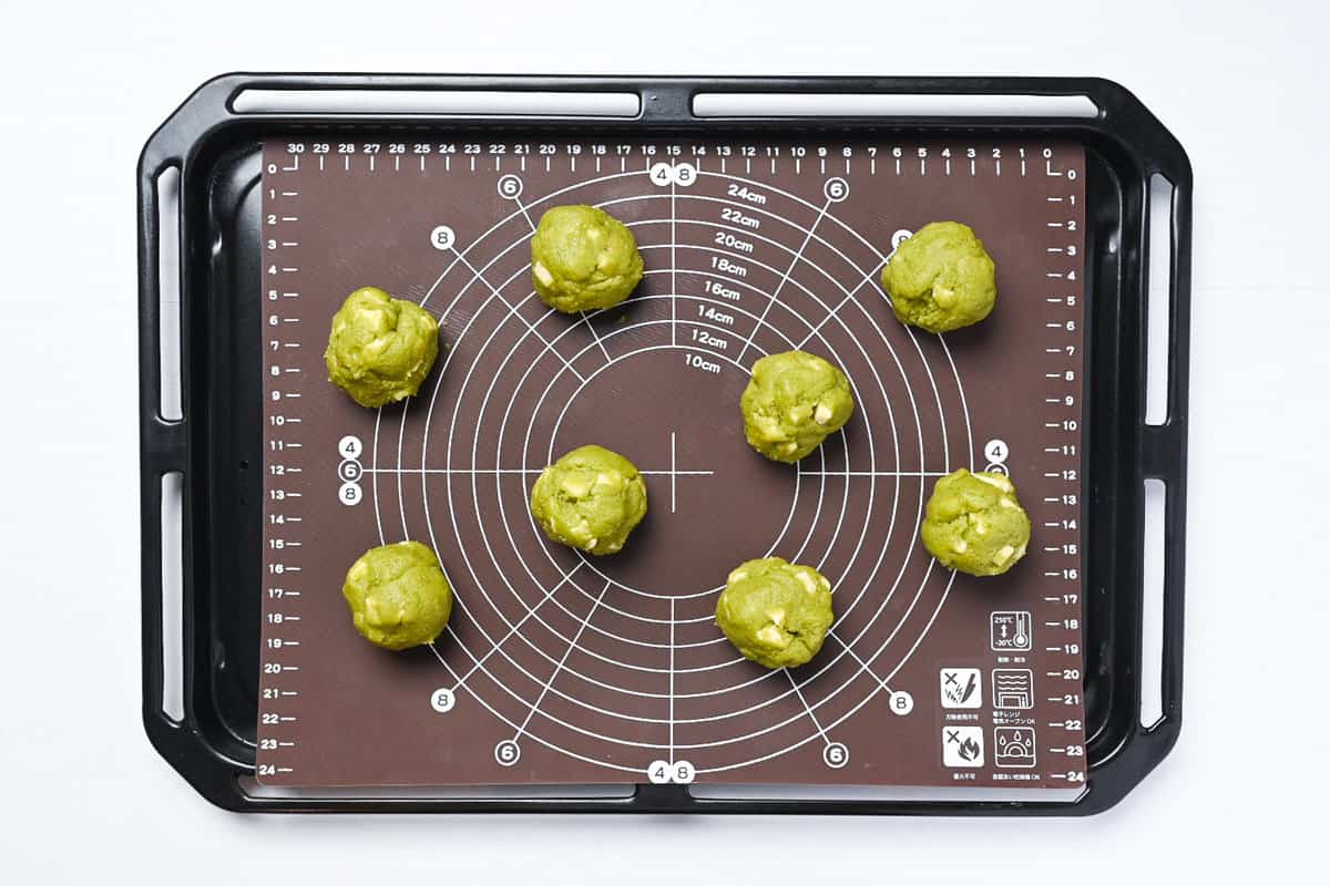 8 balls of matcha and white chocolate cookie dough laid out on a baking sheet lined with a silicone baking mat
