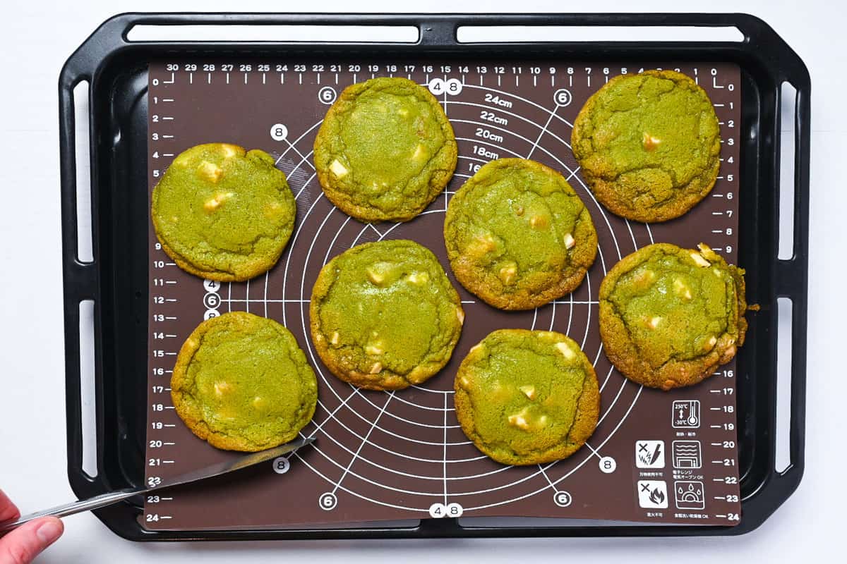 Cooked matcha and white chocolate cookies resting on a baking sheet and being shaped with a knife