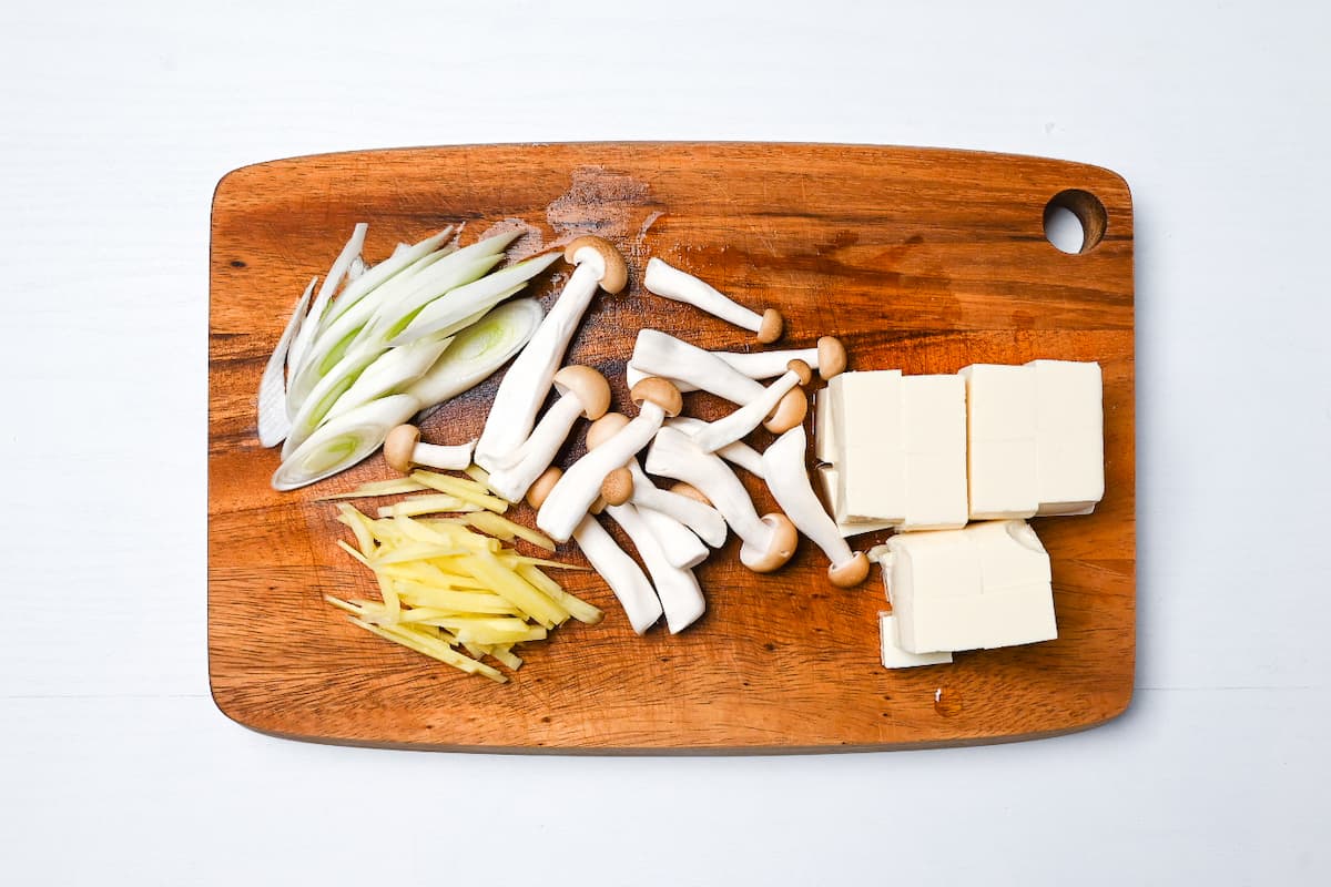 Cut spring onion, ginger, shimeji mushrooms and firm tofu on a wooden chopping board