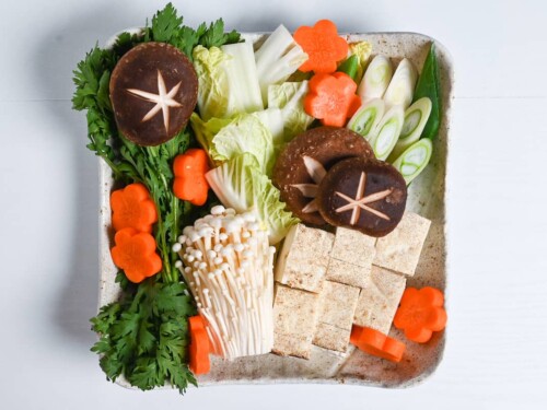 A large plate with cut vegetables and tofu for sukiyaki (includes carrots, shiitake mushrooms, enoki mushrooms, crown daisy and napa cabbage)