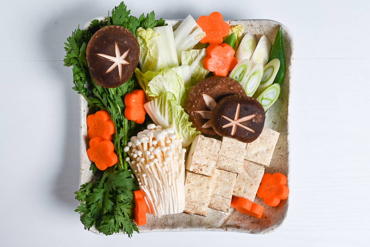 A large plate with cut vegetables and tofu for sukiyaki (includes carrots, shiitake mushrooms, enoki mushrooms, crown daisy and napa cabbage)
