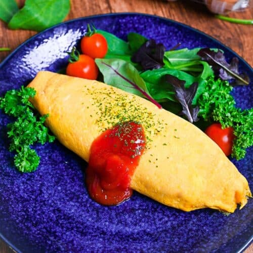 Omurice (Japanese omelette rice) topped with ketchup and served on a blue plate next to salad leaves, baby tomatoes and fresh parsley