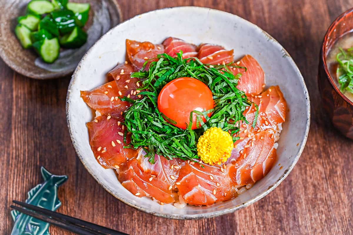 maguro zuke don (marinated tuna sashimi bowl) topped with shredded shisho, raw egg yolk and yellow flower next to pickles and miso soup