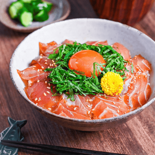 maguro zuke don (marinated tuna sashimi bowl) topped with shredded shisho, raw egg yolk and yellow flower next to pickles and miso soup