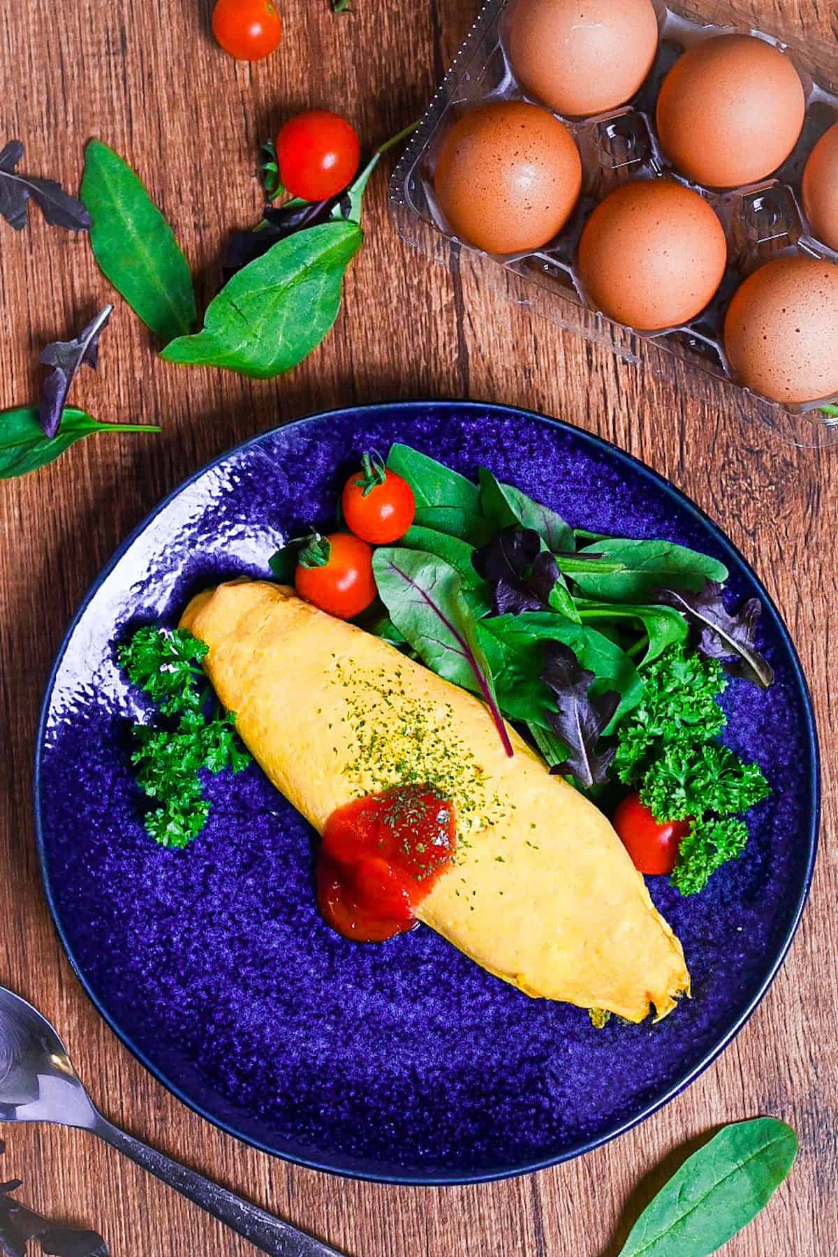 Omurice (Japanese omelette rice) topped with ketchup and served on a blue plate next to salad leaves, baby tomatoes and fresh parsley