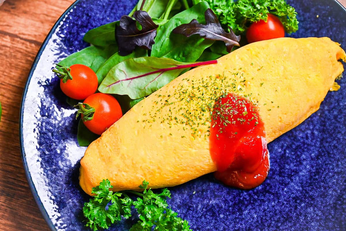 Omurice (Japanese omelette rice) topped with ketchup and served on a blue plate next to salad leaves, baby tomatoes and fresh parsley