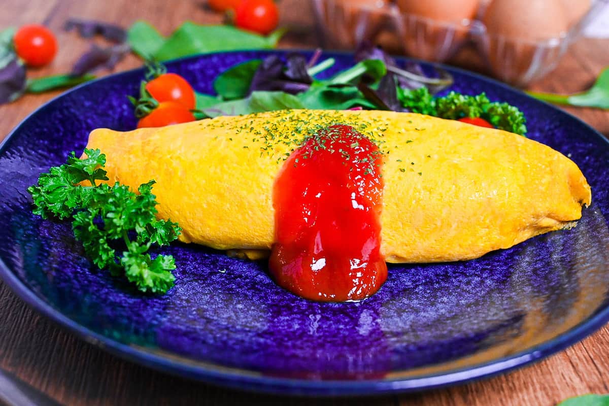 Omurice (Japanese omelette rice) topped with ketchup and served on a blue plate next to salad leaves, baby tomatoes and fresh parsley