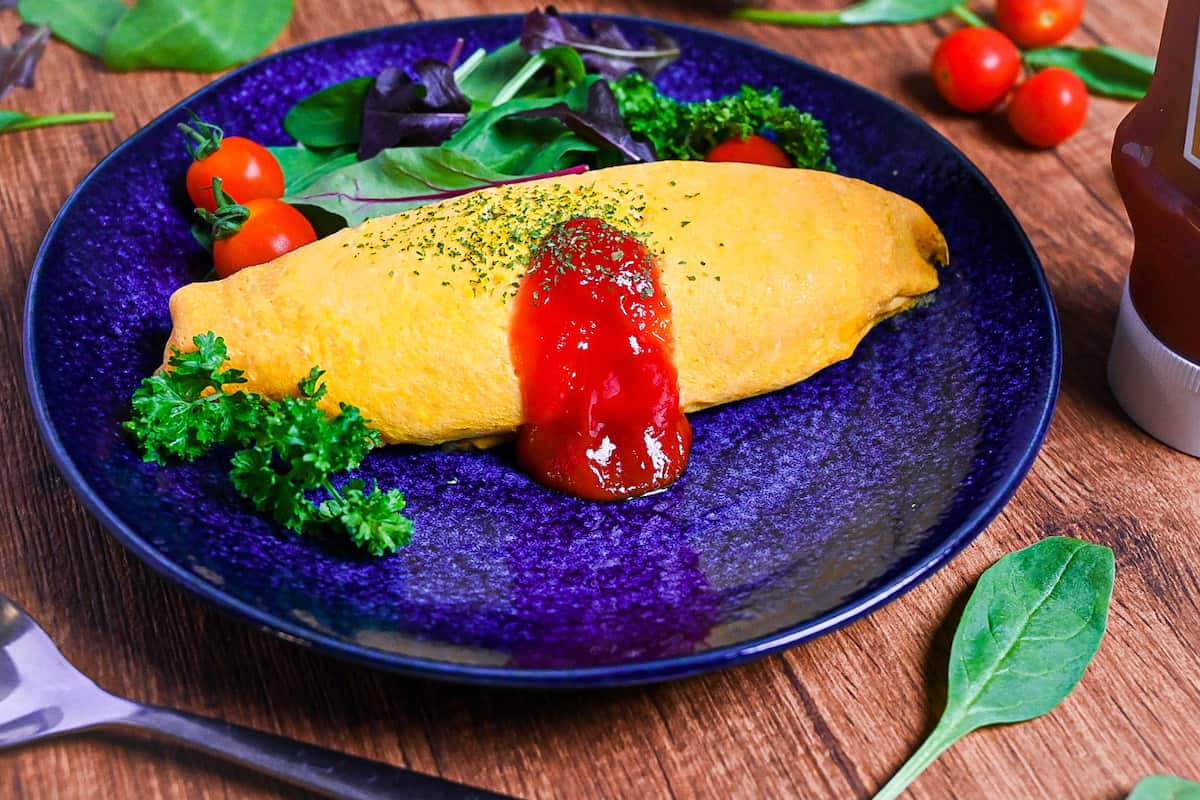 Omurice (Japanese omelette rice) topped with ketchup and served on a blue plate next to salad leaves, baby tomatoes and fresh parsley