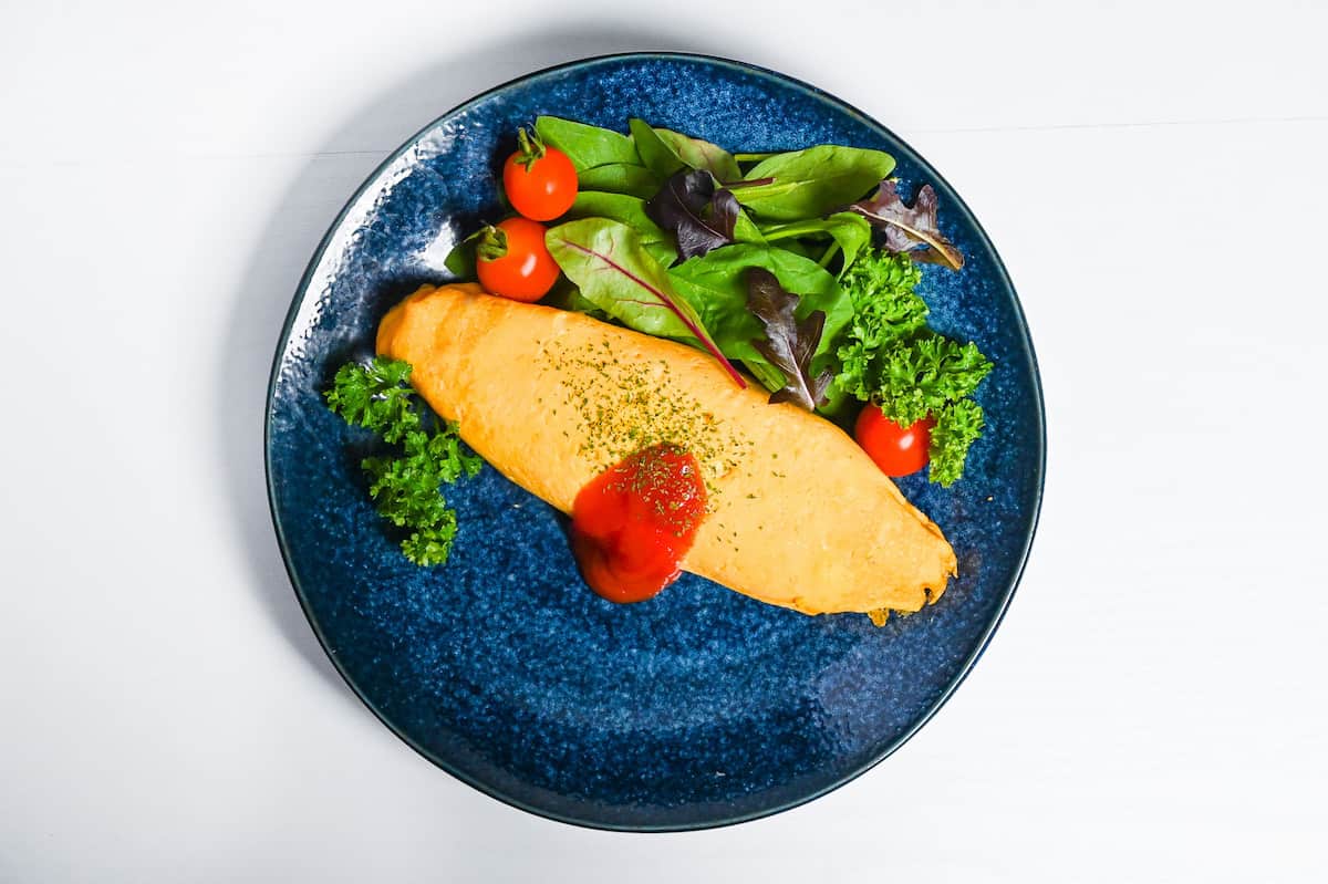 omurice on a plate topped with ketchup and dry parsley, served next to salad leaves, mini tomatoes and fresh parsley