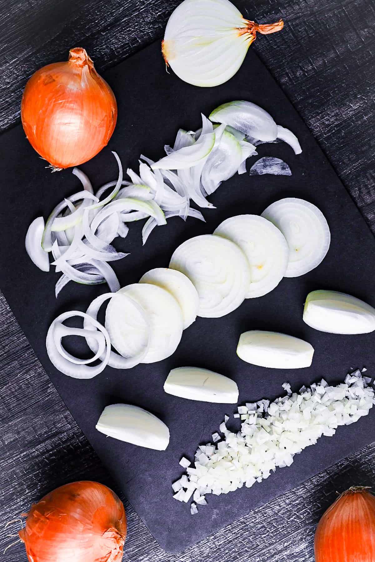 onion cut using different methods on a black chopping board - thinly sliced, thick rounds, wedges and finely diced