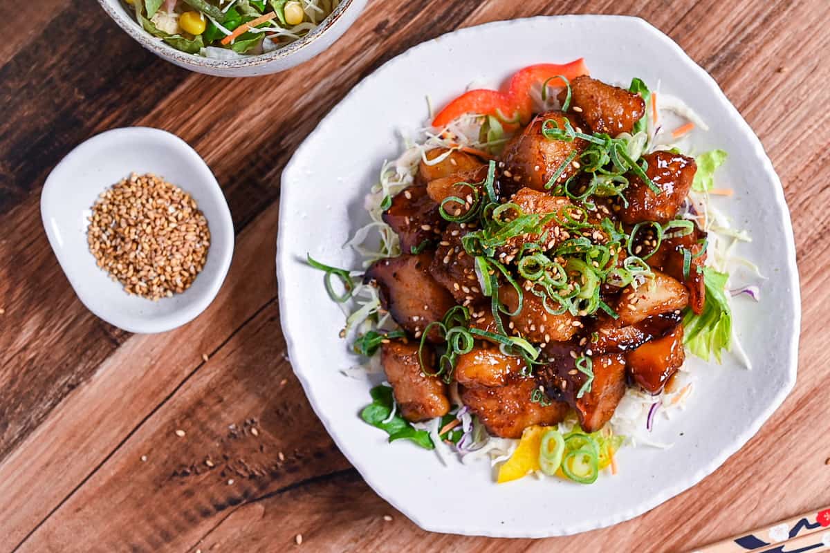 teriyaki cod served on a white plate over salad and topped with chopped green onion and toasted sesame seeds