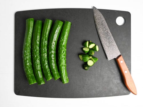 5 Japanese cucumbers with the ends cut off next to a knife on a black chopping board