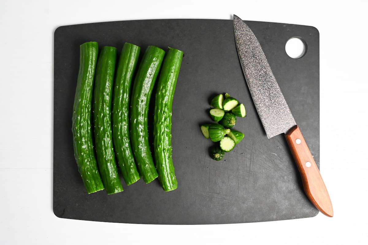 5 Japanese cucumbers with the ends cut off next to a knife on a black chopping board