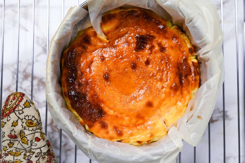 basque cheesecake in cake tin on a cooling rack with oven glove
