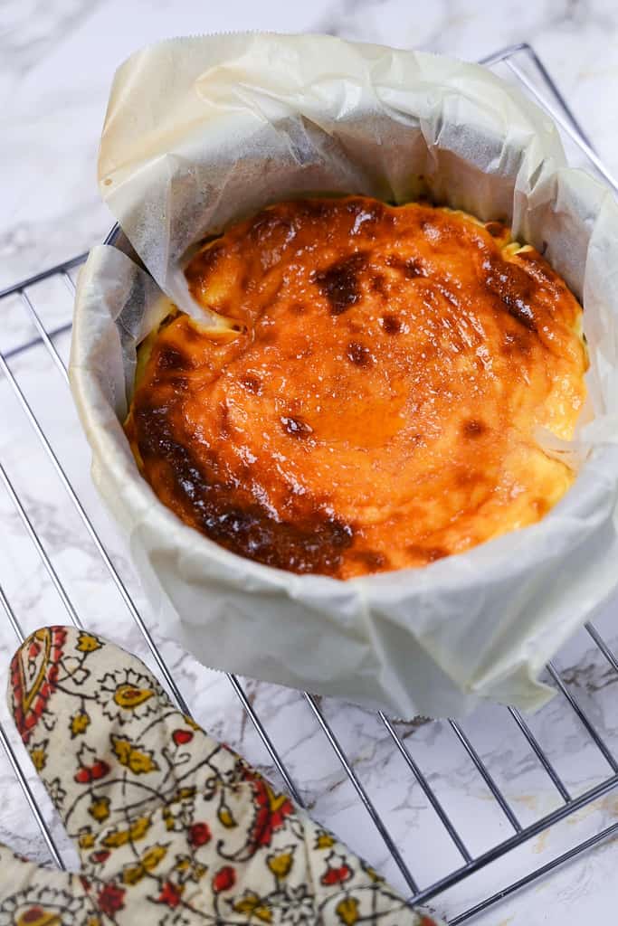 basque cheesecake in cake tin on a cooling rack with oven glove