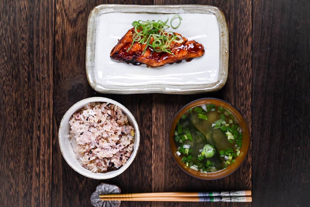 Teriyaki salmon served on a white fish plate garnished with spring onion served with multigrain rice and miso soup top down view