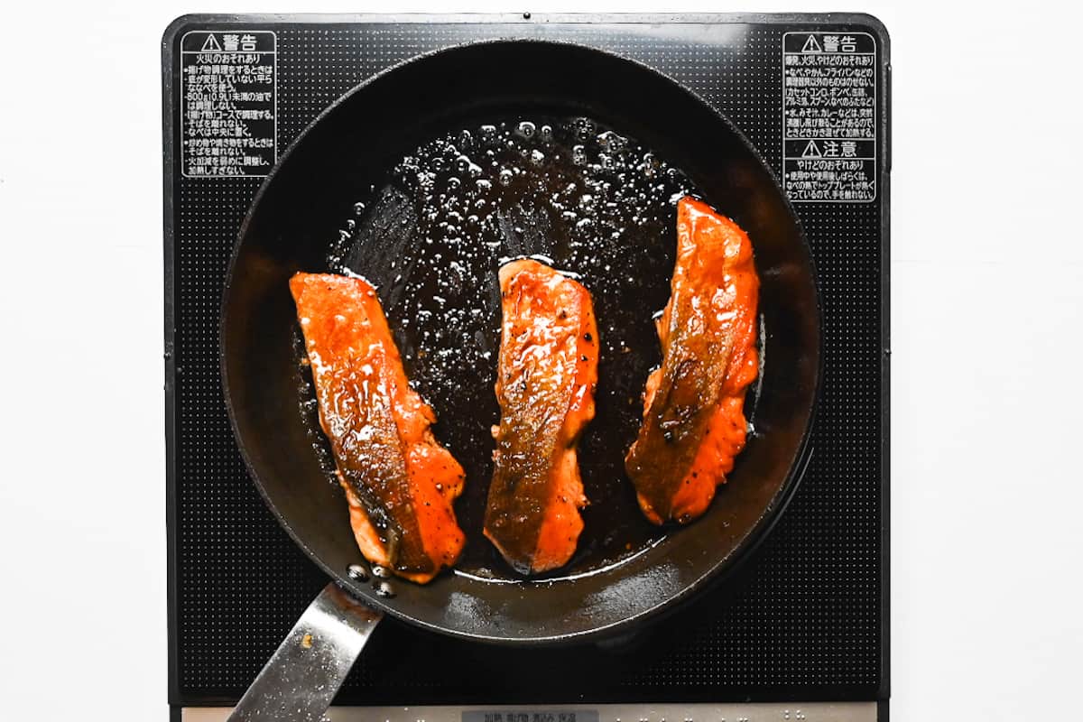 3 salmon fillets frying in a pan with teriyaki sauce