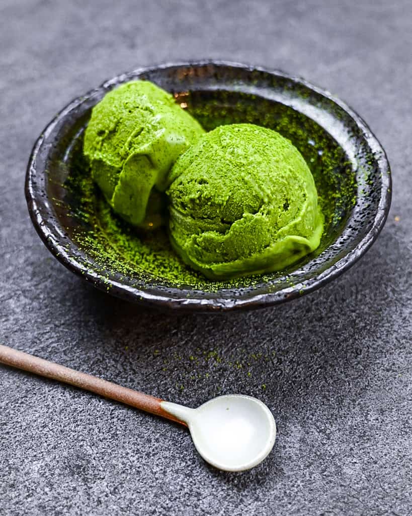 two scoops of matcha green tea ice cream served in a black bowl with ceramic spoon