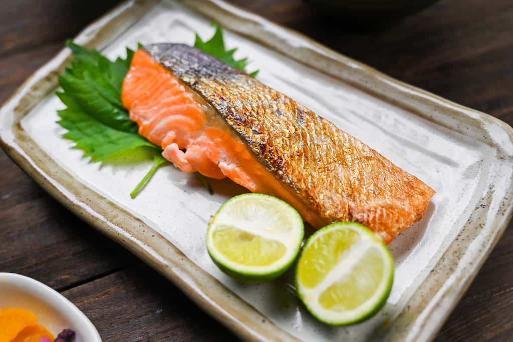 Shizake Japanese Salted Salmon served on a pale rectangular plate with sudachi and shiso leaves left side view