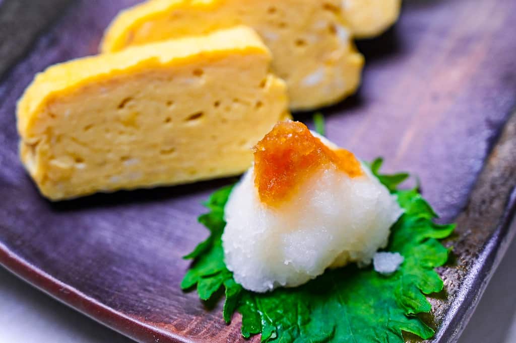 Pieces of sliced dashimaki tamago served on a brown plate with grated daikon