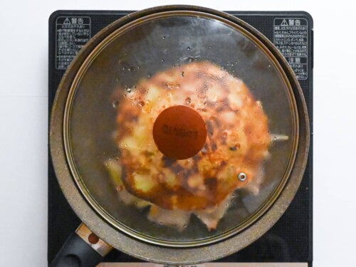 Flipped Hiroshima style okonomiyaki frying in a frying pan with a lid