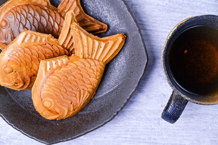 Three golden Taiyaki (fish shaped red bean cake) on a brown plate with cup of tea