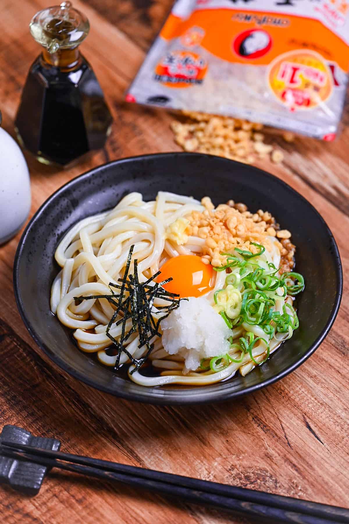 Chilled bukkake udon in a black dish topped with tempura bits, chopped green onion, grated daikon, shredded nori and an egg yolk