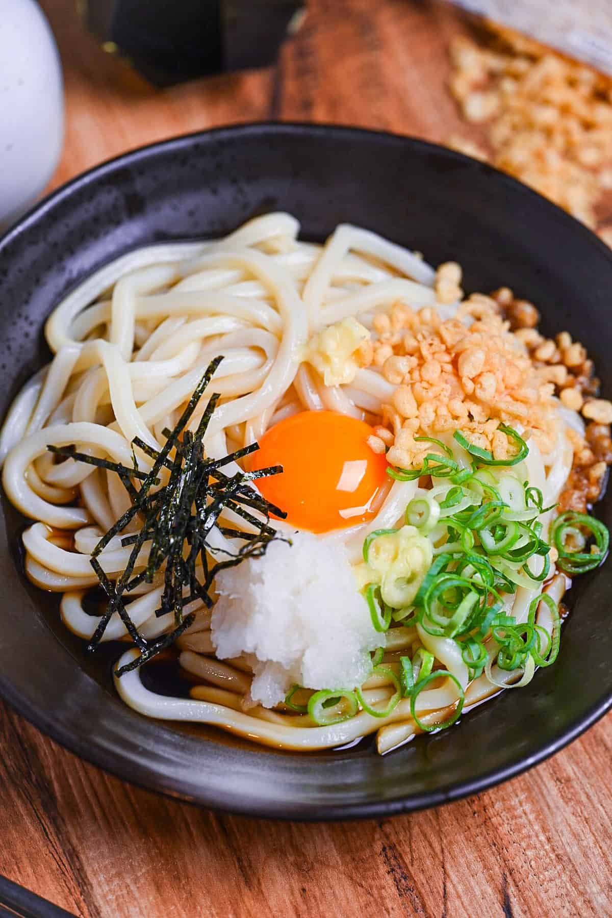 Chilled bukkake udon in a black dish topped with tempura bits, chopped green onion, grated daikon, shredded nori and an egg yolk close-up vertical