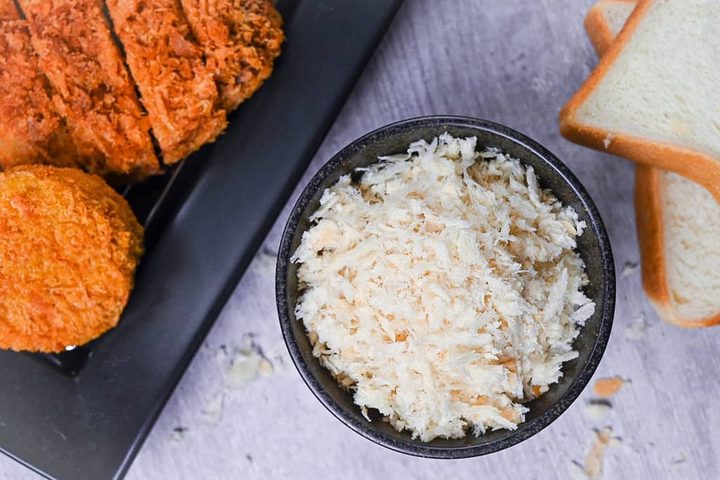 Japanese panko breadcrumbs in a black bowl next to a tray of tonkatsu and croquettes and 2 slices of Japanese white bread (shokupan)