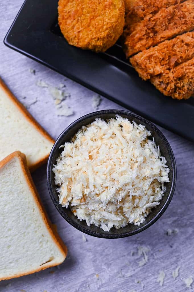 Japanese panko breadcrumbs in a black bowl next to a tray of tonkatsu and croquettes and 2 slices of Japanese white bread (shokupan)