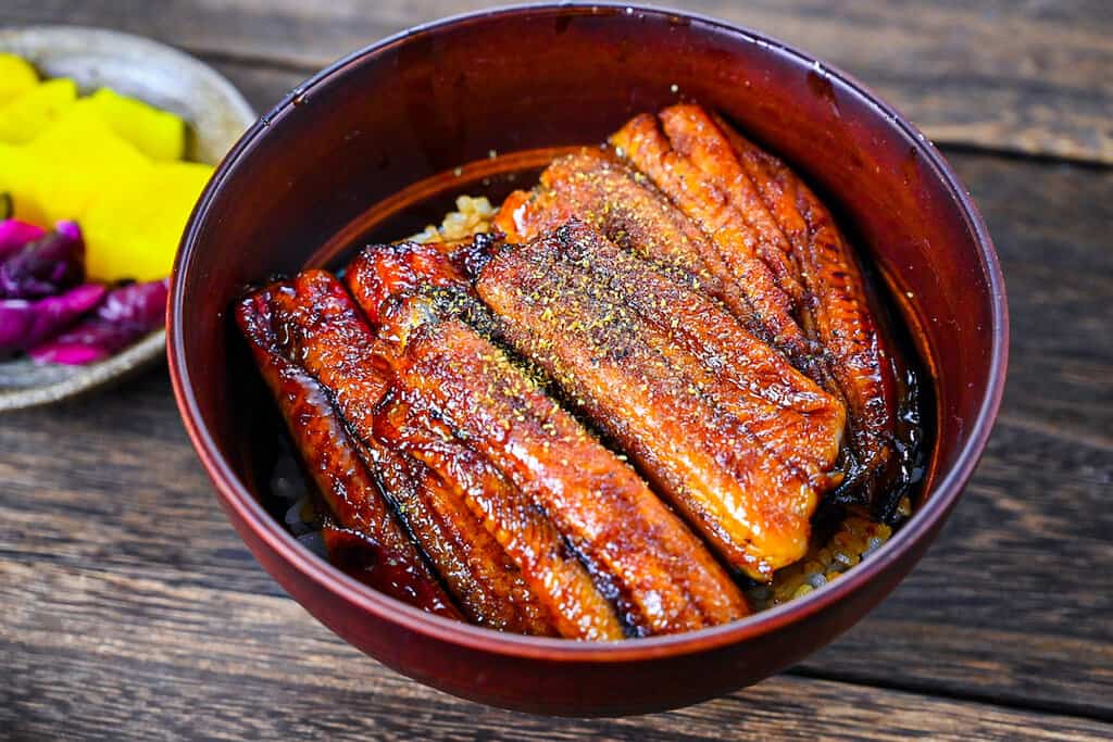 Unagi don (Japanese Grilled Eel Rice Bowl) sprinkled with sansho Japanese pepper and pickles on the side
