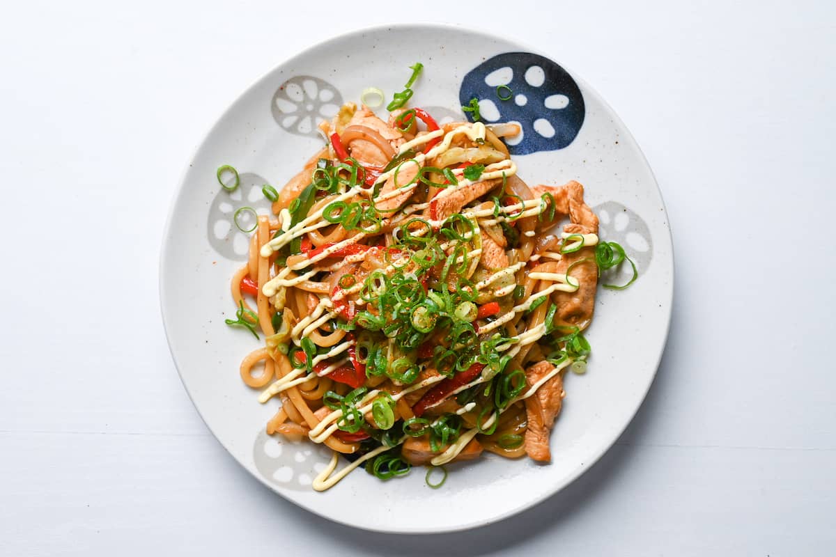Chicken yaki udon flavoured with garlic, butter and soy sauce on a white plate drizzled with Japanese mayo and sprinkled with chopped spring onion