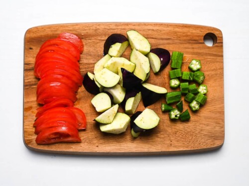 Sliced tomato and chopped eggplant and okra on a wooden chopping board