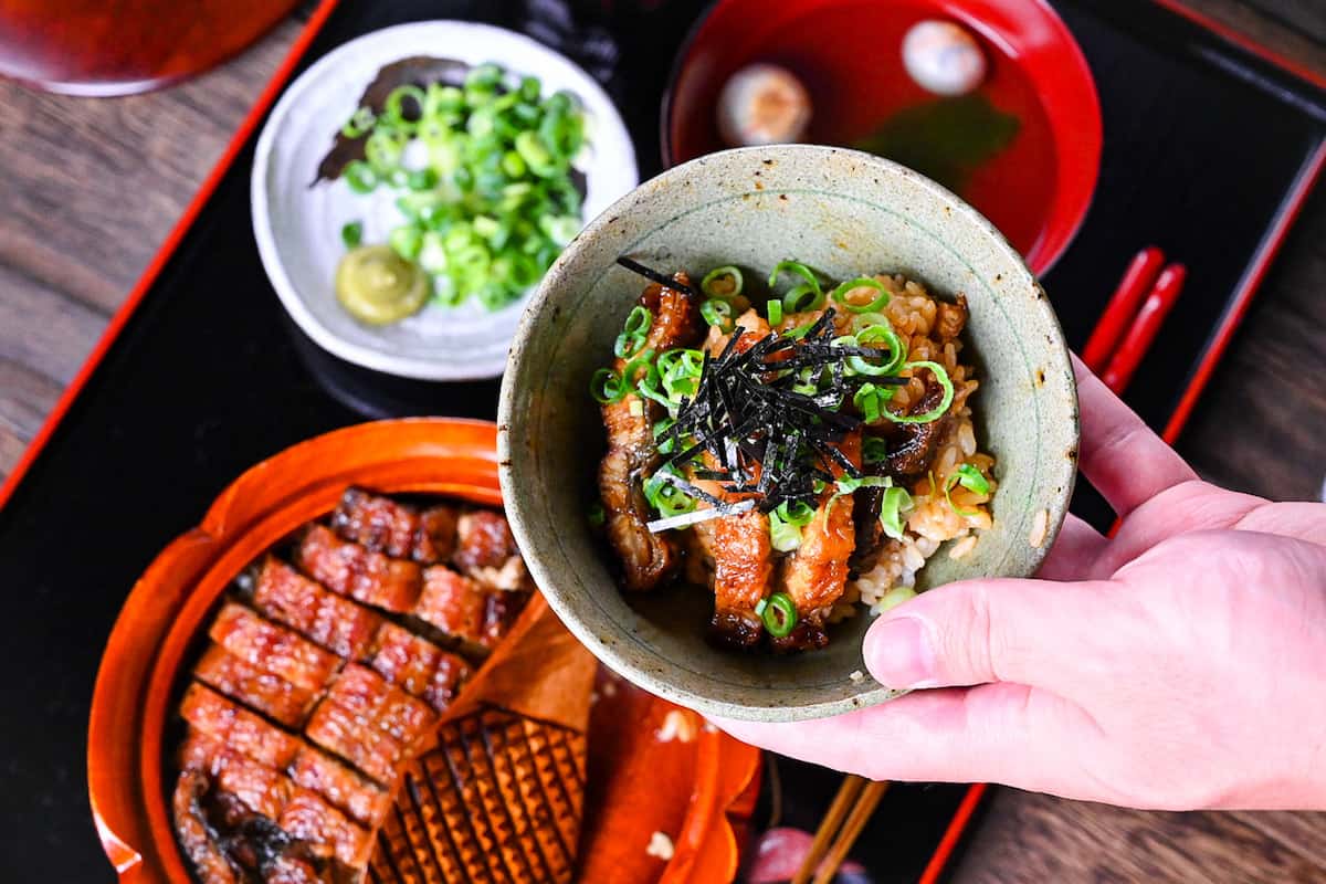 A portion of Hitsumabushi in a green rice bowl with toppings and soup in the background