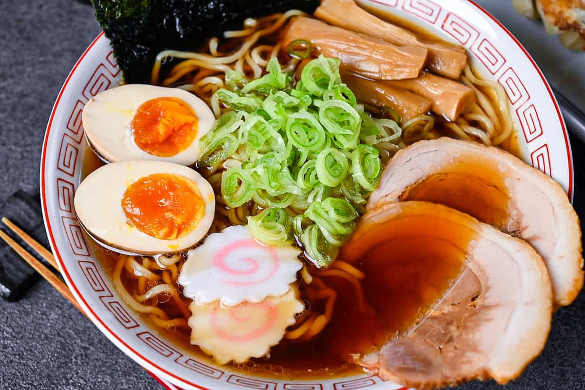 Homemade shoyu ramen topped with ramen egg, menma, narutomaki, chashu, chopped spring onions and nori in a white and red Chinese style noodle bowl