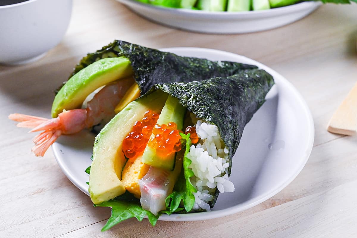two temaki sushi hand rolls on a white plate with ingredients in the background