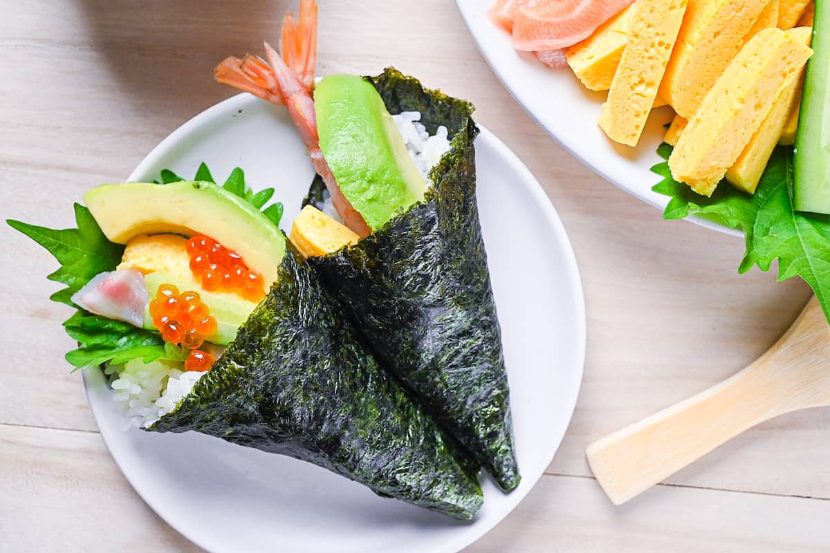 two temaki sushi hand rolls on a white plate with ingredients on the side