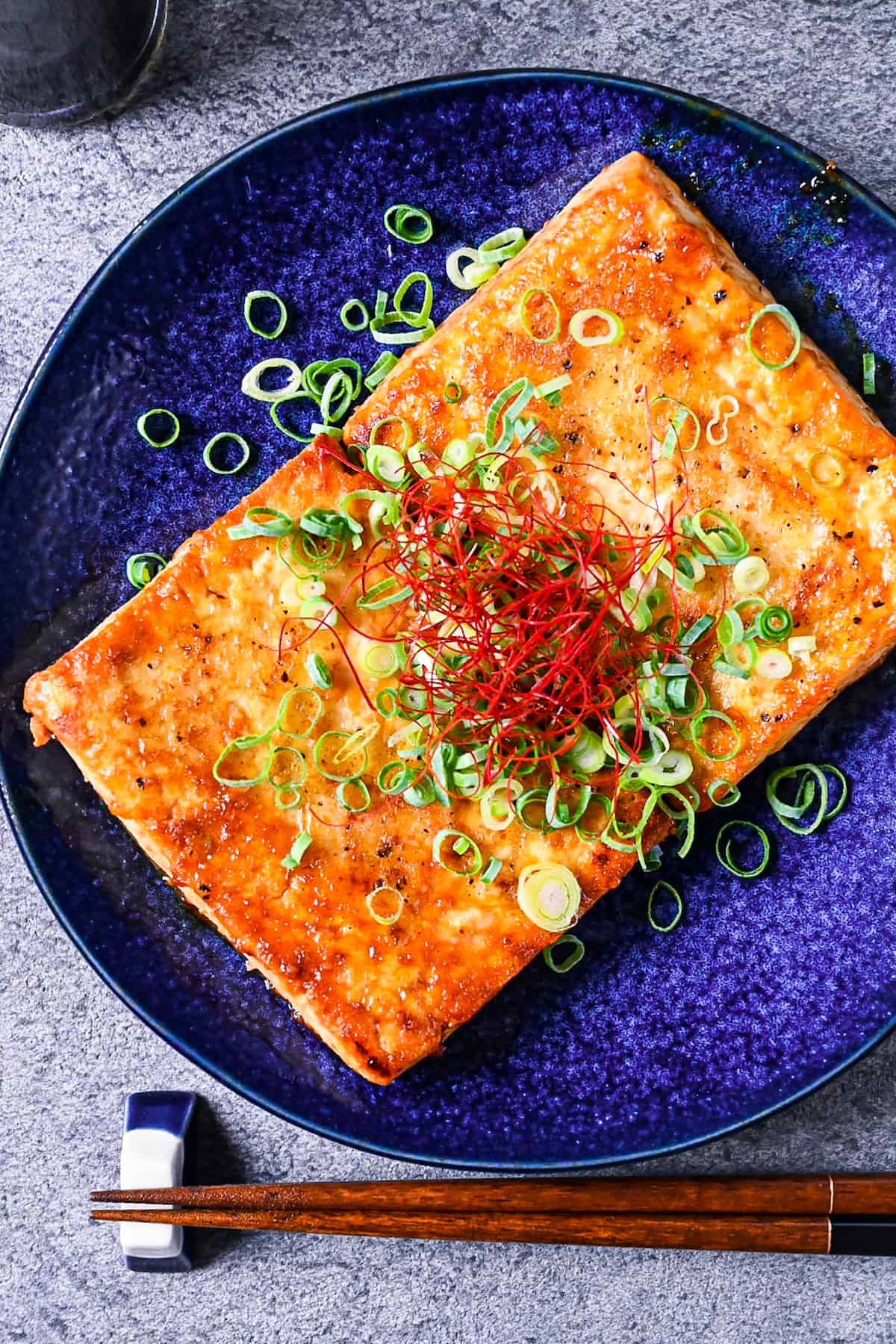 Tofu steak coated in a Japanese style sauce topped with spring onion and chili threads on a blue plate top down view