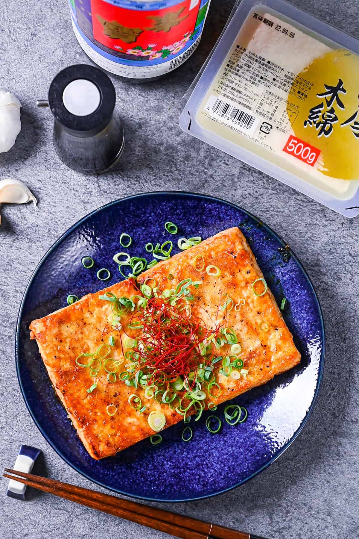 Tofu steak coated in a Japanese style sauce topped with spring onion and chili threads on a blue plate with ingredients of tofu, soy sauce, mirin and garlic