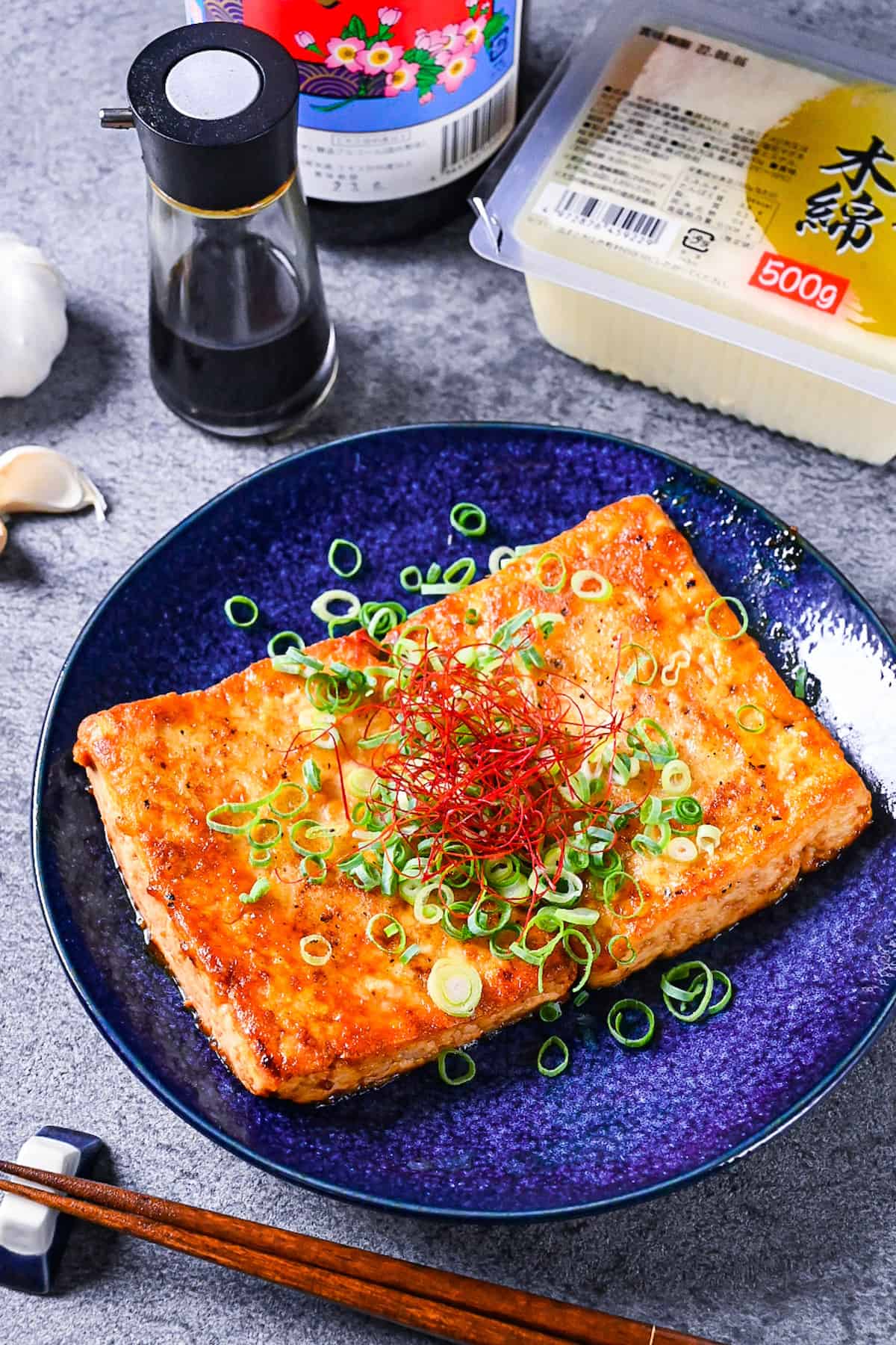 Tofu steak coated in a Japanese style sauce topped with spring onion and chili threads on a blue plate with ingredients of tofu, soy sauce, mirin and garlic