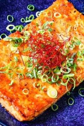 Tofu steak coated in a Japanese style sauce topped with spring onion and chili threads on a blue plate thumbnail