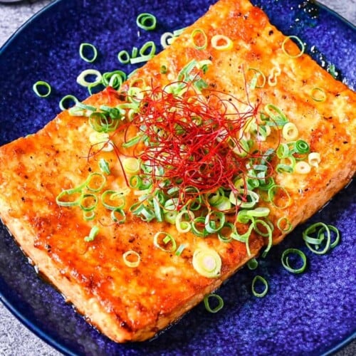 Tofu steak coated in a Japanese style sauce topped with spring onion and chili threads on a blue plate thumbnail