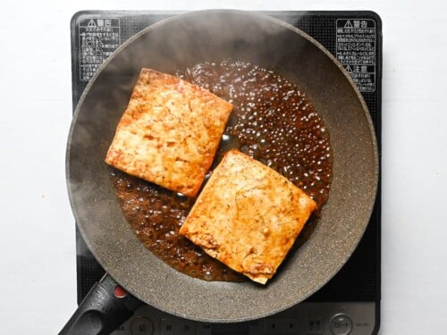 Two pieces of tofu in a frying pan with bubbling Japanese style sauce