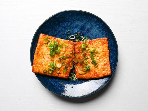 Two tofu steaks on a blue plate garnished with spring onion and red chili threads